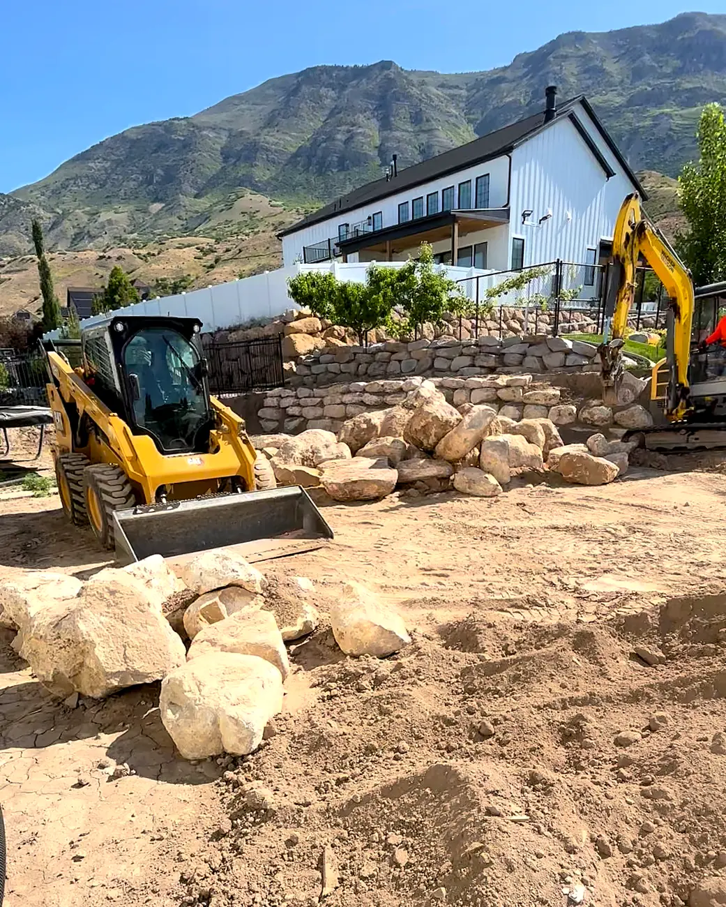 Skid steer and excavator building a boulder wall in a backyard below the mountains