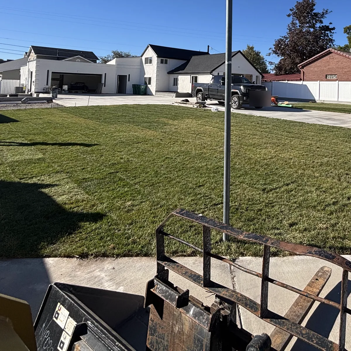 Freshly laid sod on a corner lot in front of a white home, with skid steer forks in the foreground
