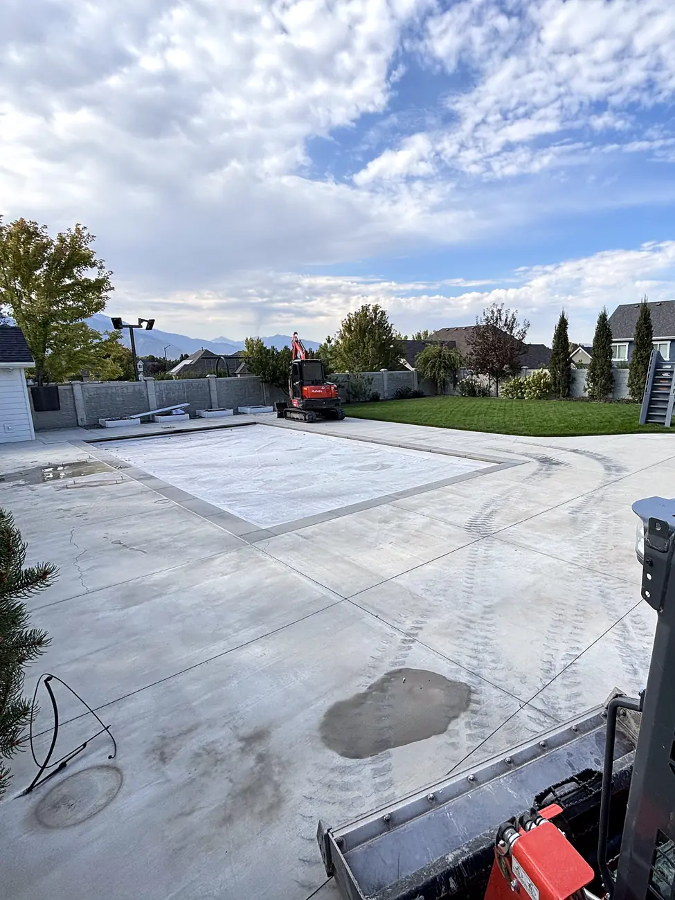 Large concrete pool deck in a backyard before removal, with a skid steer ready to start