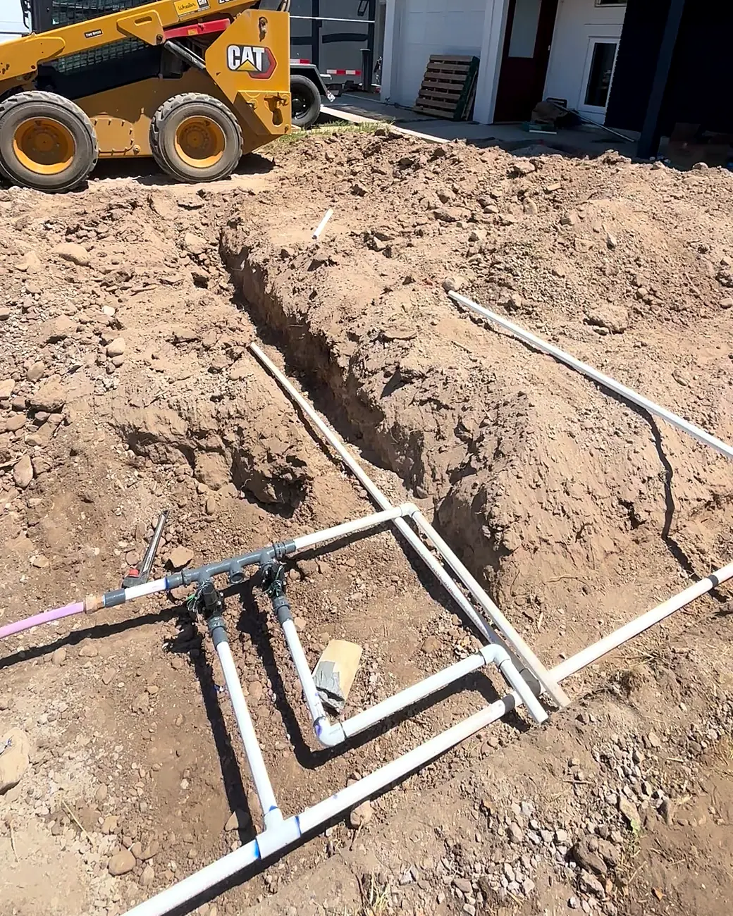 New sprinkler valves and pipe in open trenches at a new build, with a skid steer behind